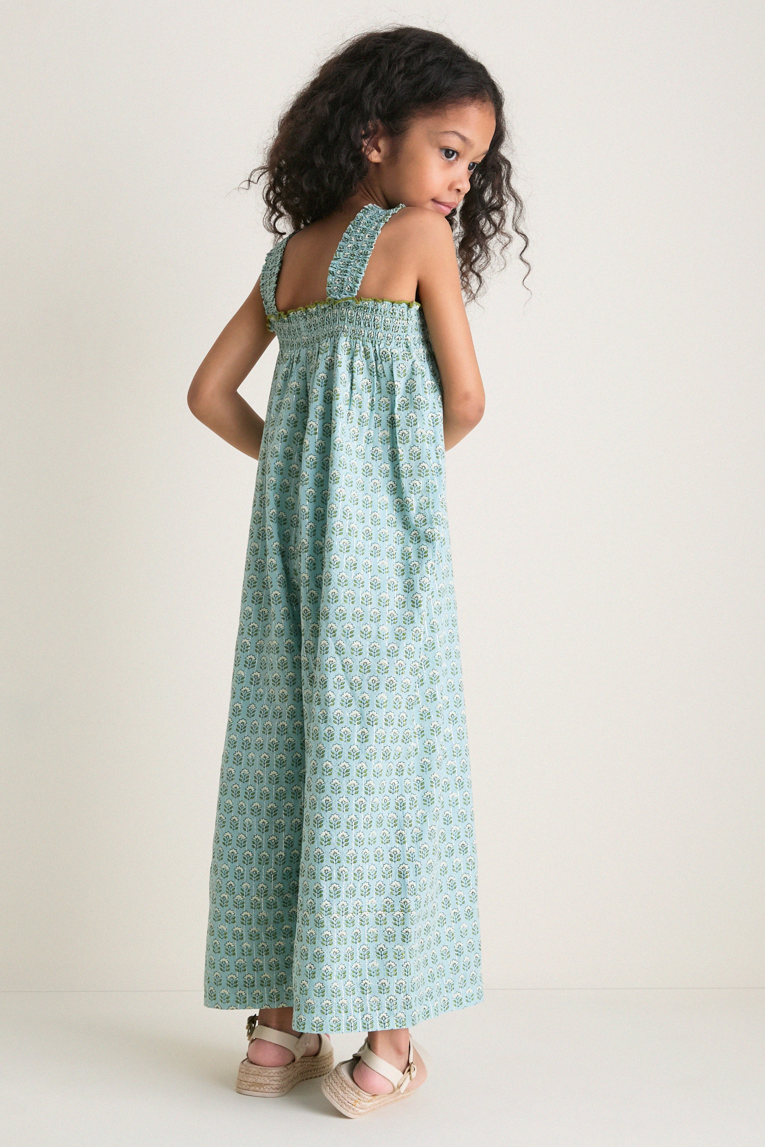 Young girl in a classic, teal-green Blue Prairie Block Print Eleven Jumper, showcasing effortless style with smocked bodice and wide-leg design, paired with beige espadrilles.
