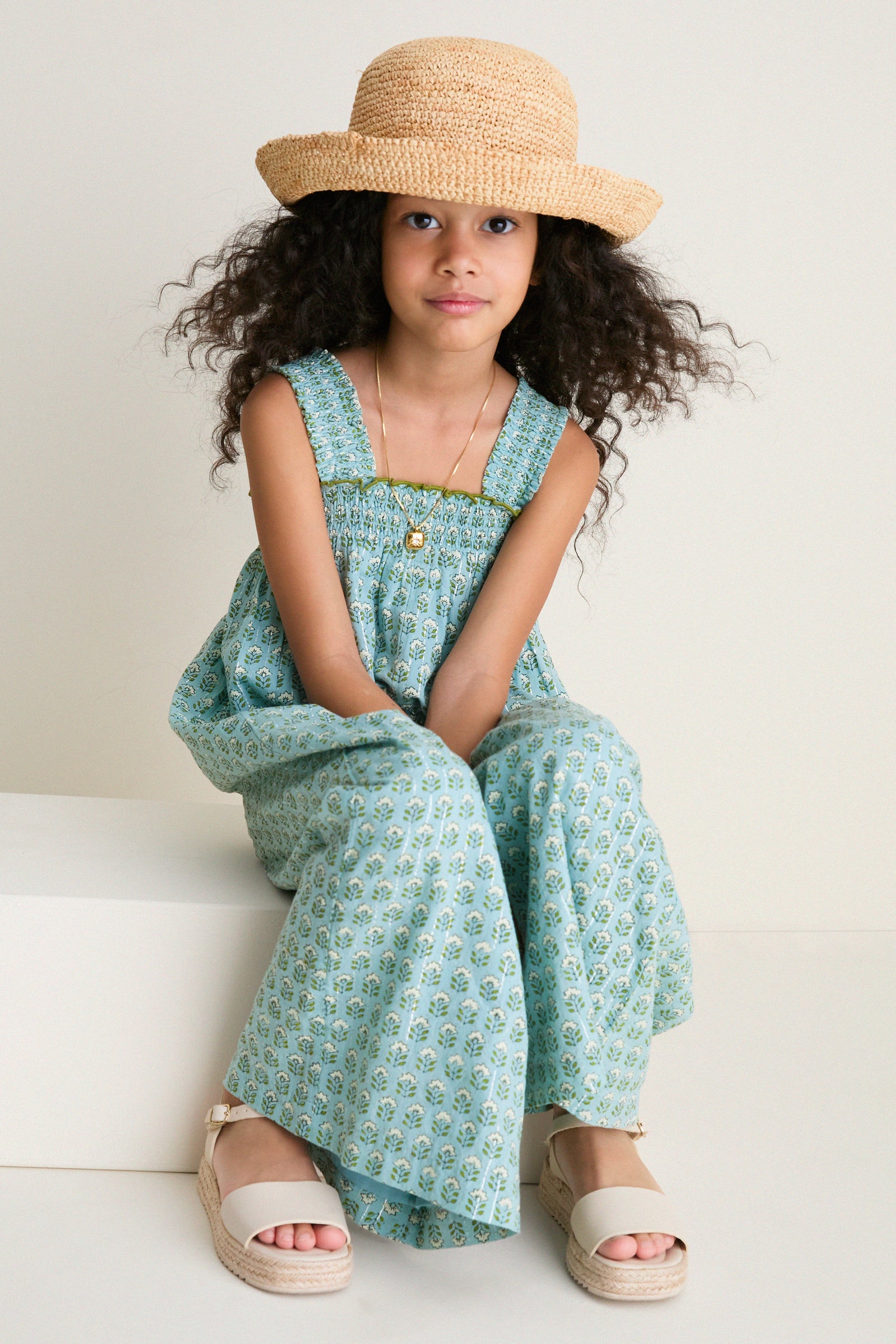 Young girl in a classic Blue Prairie Block Print Eleven Jumper with floral motifs, smocked bodice, and espadrille sandals, exuding effortless charm and high-end playfulness.