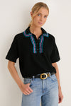 Woman models Deep Black Multi Polo Sweater, a classic yet fun knit with teal and purple scalloped trim, paired effortlessly with light-wash jeans and a gold-buckled belt.