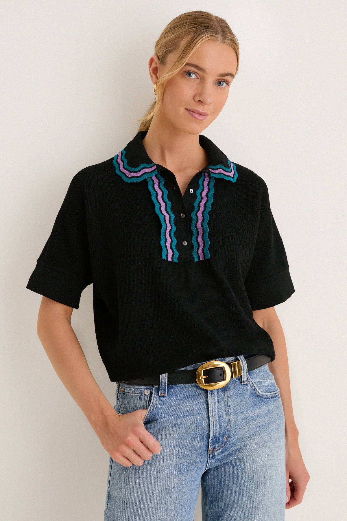Woman models Deep Black Multi Polo Sweater, a classic yet fun knit with teal and purple scalloped trim, paired effortlessly with light-wash jeans and a gold-buckled belt.