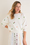 Young woman models the classic Milk Multi Dump Sweater, featuring effortless, fun embroidery of clovers, ladybugs, and butterflies, paired with a matching high-end knit skirt.