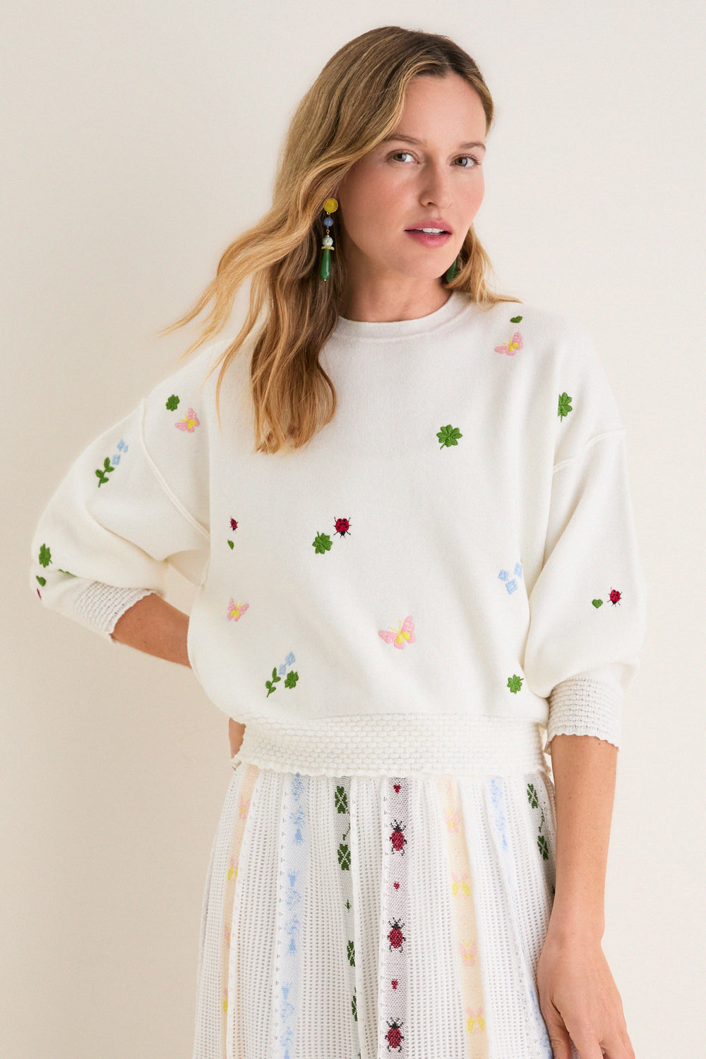 Young woman models the classic Milk Multi Dump Sweater, featuring effortless, fun embroidery of clovers, ladybugs, and butterflies, paired with a matching high-end knit skirt.