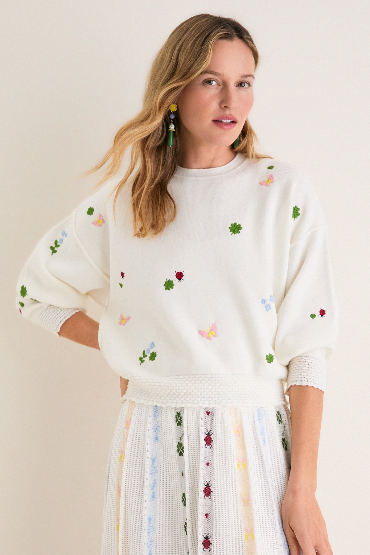 Young woman models the classic Milk Multi Dump Sweater, featuring effortless, fun embroidery of clovers, ladybugs, and butterflies, paired with a matching high-end knit skirt.