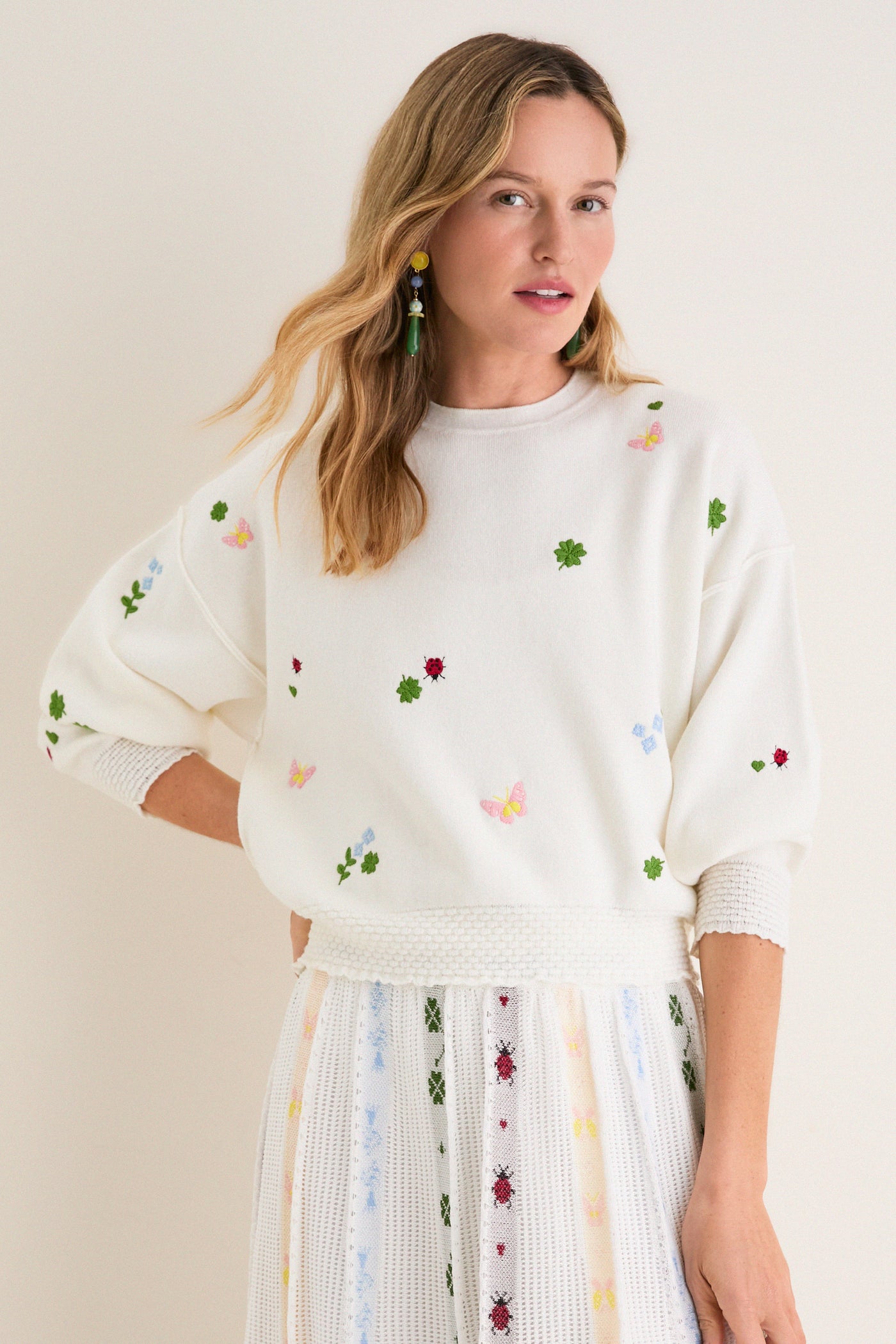Young woman models the classic Milk Multi Dump Sweater, featuring effortless, fun embroidery of clovers, ladybugs, and butterflies, paired with a matching high-end knit skirt.