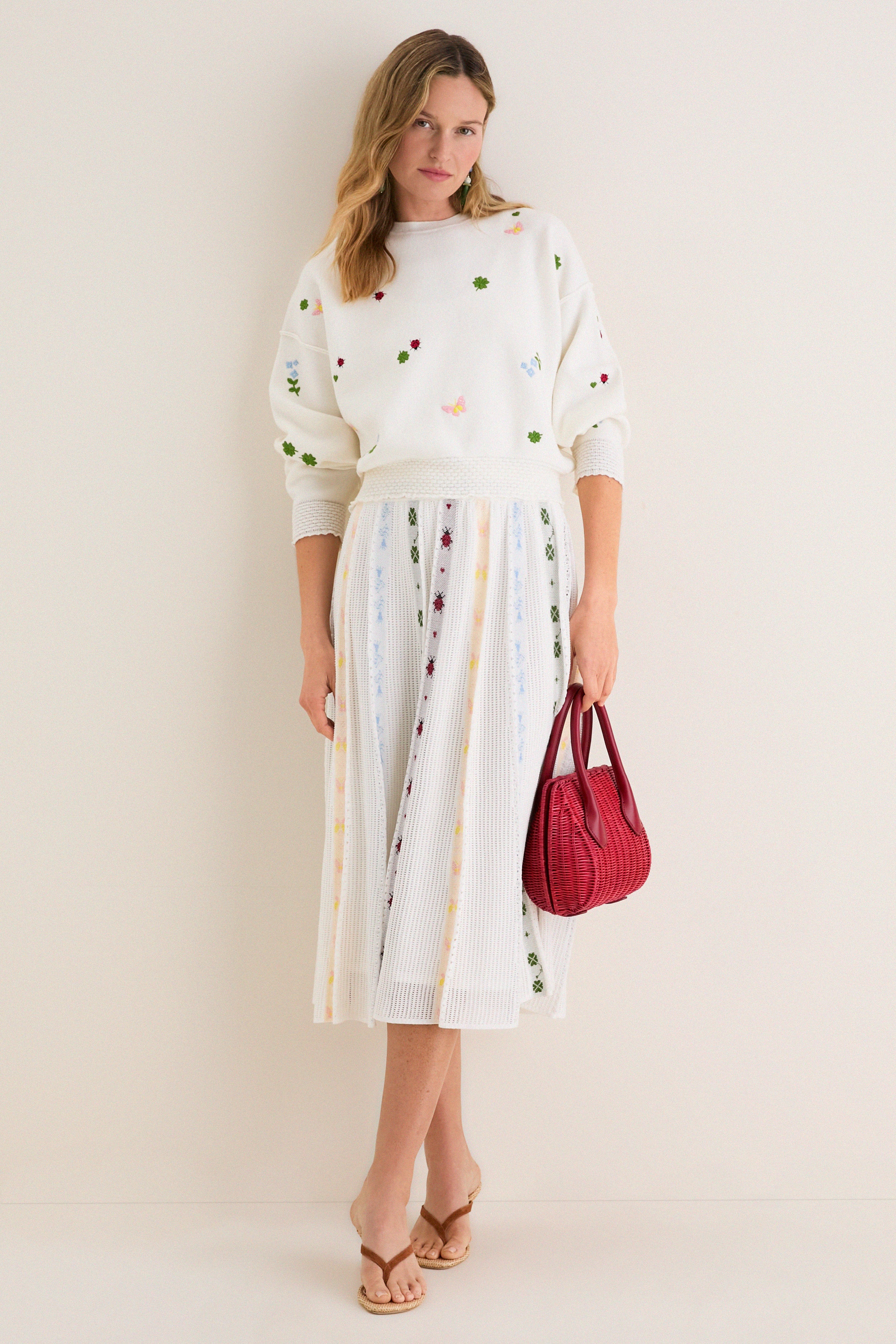 Woman models Milk Multi Dump Sweater with floral embroidery and smocked cuffs, styled effortlessly with a matching skirt, exuding a classic and fun high-end spring look.
