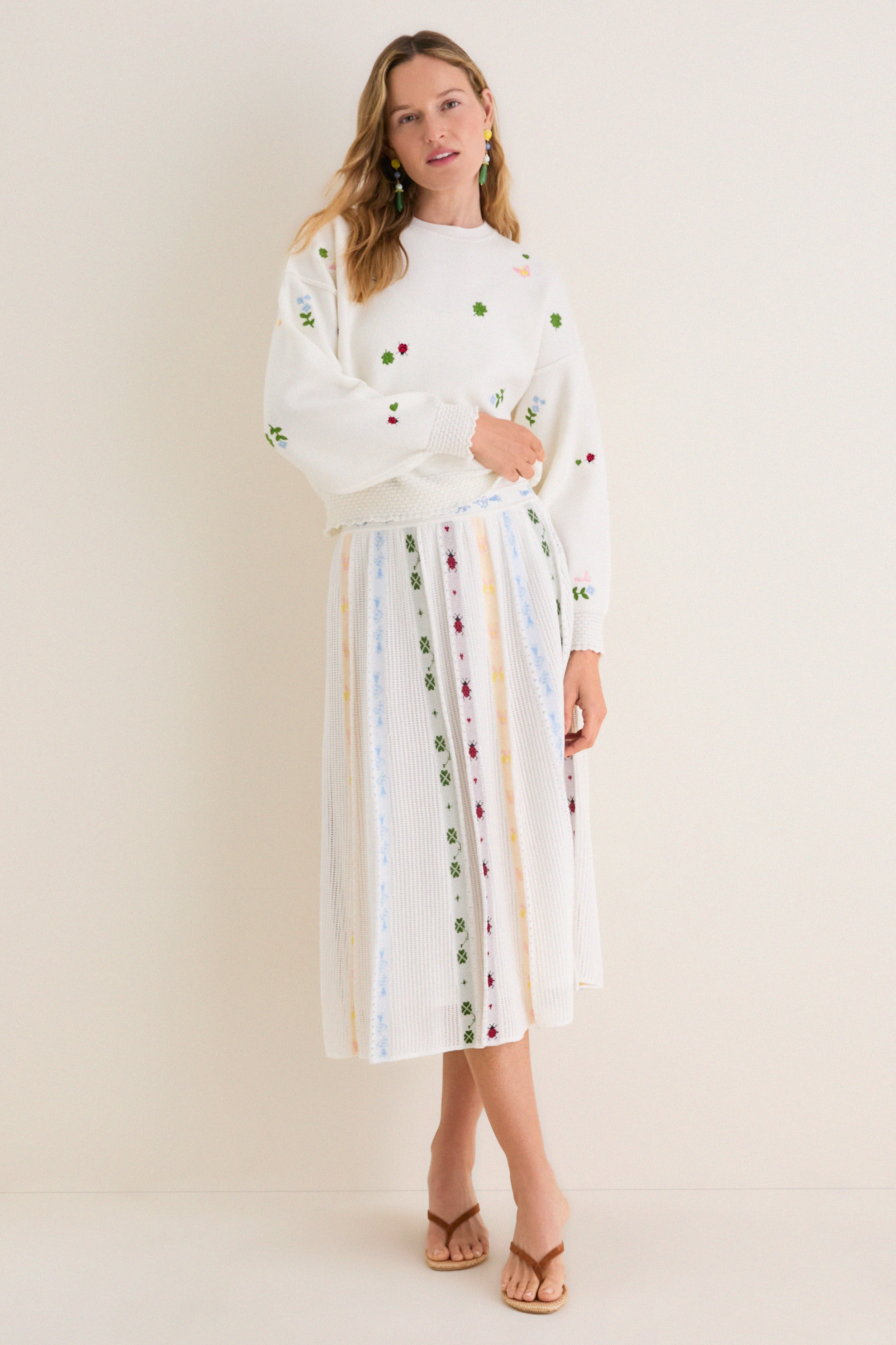 Woman in a classic, embroidered Milk Multi Dump Sweater and matching skirt, exuding an effortless, fun, high-end style with colorful earrings and brown sandals.