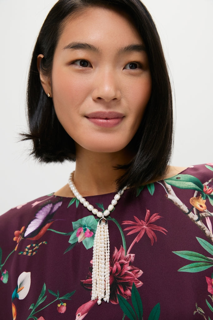 Woman wearing the Pearl Delfina Necklace, featuring large pearls and a multi-strand tassel pendant, paired with a jewel-tone, floral-and-bird patterned blouse.