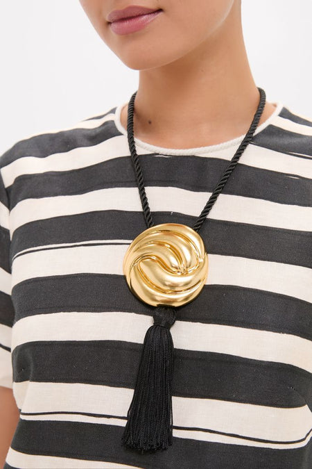 Person wearing the Gold and Black Hebe Necklace, featuring a classic gold-tone medallion with a black tassel on a thick cord, exuding effortless, fun, high-end style.