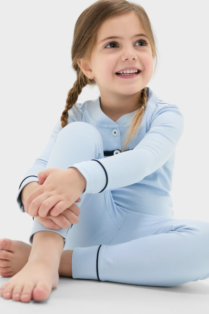 Young girl in a classic, effortless Light Blue Pima Cotton Arien Kid's Pajama Set, featuring fun contrast trim and mandarin collar, smiling while sitting barefoot on the floor.