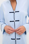 Close-up of hands fastening the Light Blue Wilford Women's Pajama Set, featuring a classic mandarin collar, effortless black piping, and high-end frog-style button closures.