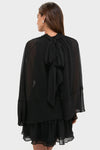 Woman in a classic, effortless Black Cape Chiffon Mayfield Dress with sheer cape sleeves and a fun, high-end layered skirt, tied elegantly at the back.