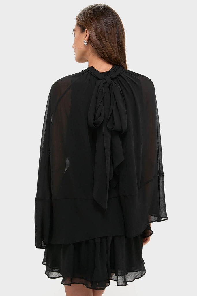 Woman in a classic, effortless Black Cape Chiffon Mayfield Dress with sheer cape sleeves and a fun, high-end layered skirt, tied elegantly at the back.