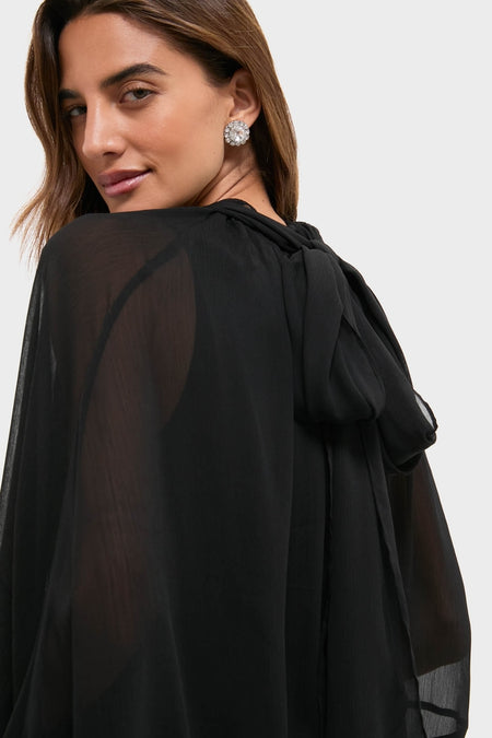 Woman in Black Cape Chiffon Mayfield Dress with sheer long sleeves and back bow, showcasing an effortless, classic style with a fun, high-end appeal.