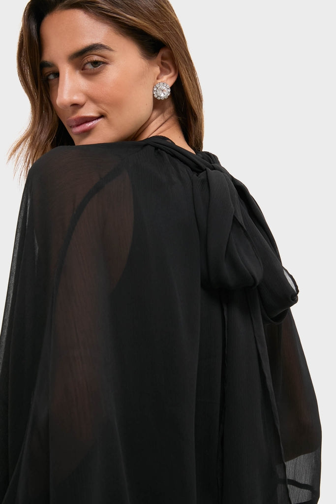 Woman in Black Cape Chiffon Mayfield Dress with sheer long sleeves and back bow, showcasing an effortless, classic style with a fun, high-end appeal.