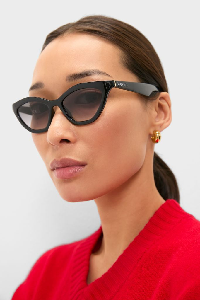 Woman wearing Shiny Black Cat-Eye Mods Sunglasses with Gucci logo, styled with gold hoop earrings and a red knit top, embodying a classic, effortless, and high-end fashion look.