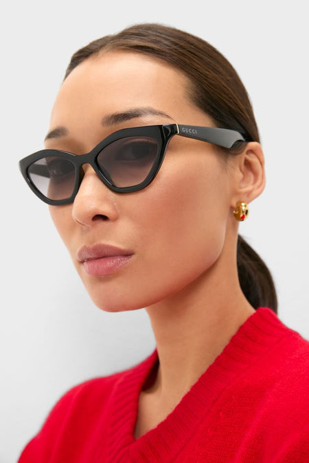 Woman wearing Shiny Black Cat-Eye Mods Sunglasses with Gucci logo, styled with gold hoop earrings and a red knit top, embodying a classic, effortless, and high-end fashion look.