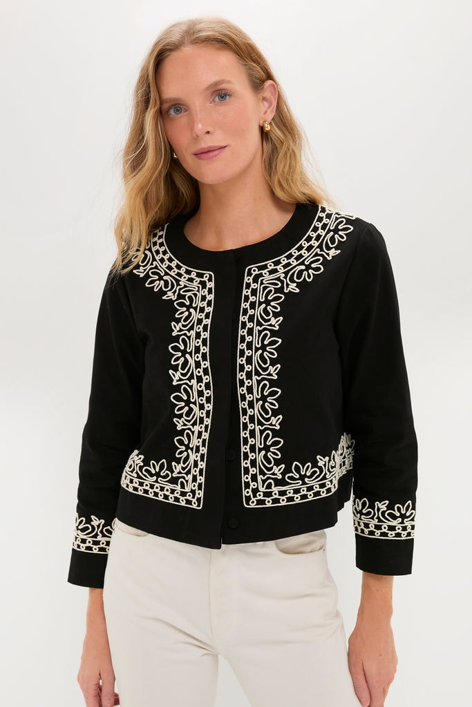 Woman models the classic Soutache Cotton Maddy Jacket, featuring effortless white floral embroidery on black fabric, paired with chic off-white pants for a fun, high-end look.