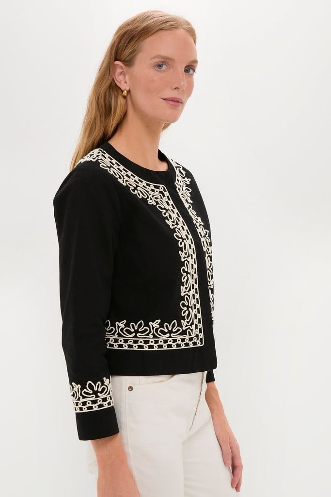Woman wearing the classic Soutache Cotton Maddy Jacket with intricate white embroidery, paired with cream trousers, reflecting an effortless, fun, high-end style.