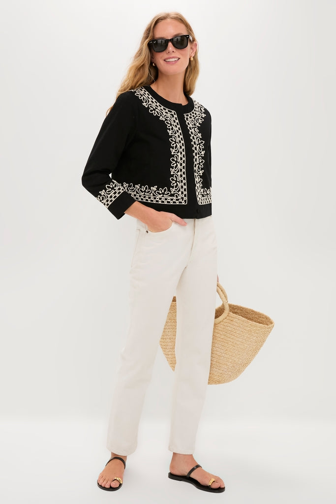 Woman wearing a classic, effortless Soutache Cotton Maddy Jacket with floral embroidery, paired with high-waisted jeans and sandals, holding a straw tote for a fun, high-end look.