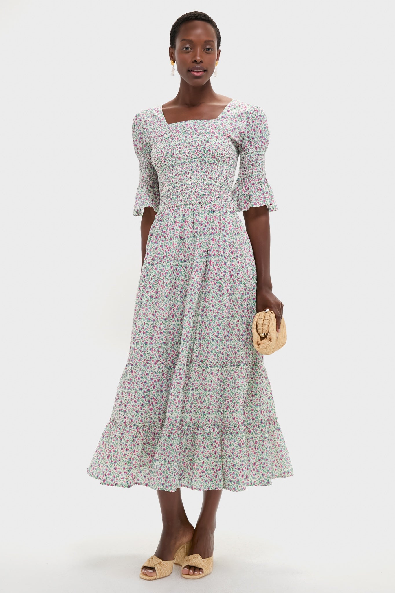 Primrose Print Bea Dress: Woman in a classic, effortless midi sundress with a floral pattern, puff sleeves, and tiered skirt, accessorized with a straw clutch and woven mules.