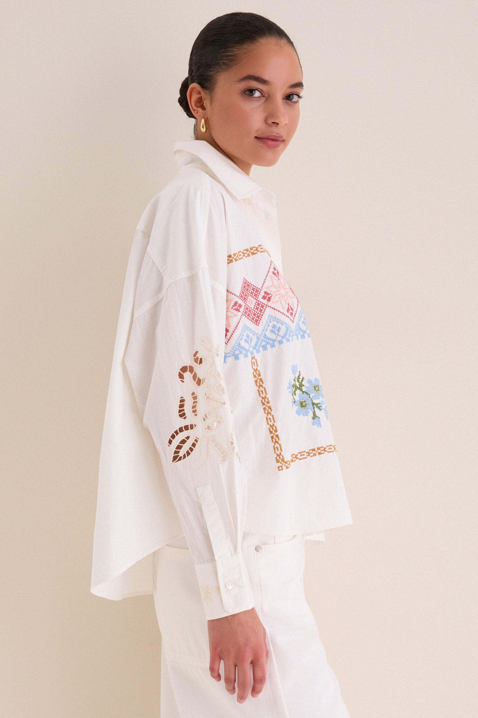 Model in profile wearing the White Fonte Shirt, a classic, effortlessly chic blouse with colorful cross-stitch embroidery and eyelet lace, embodying high-end, fun style.
