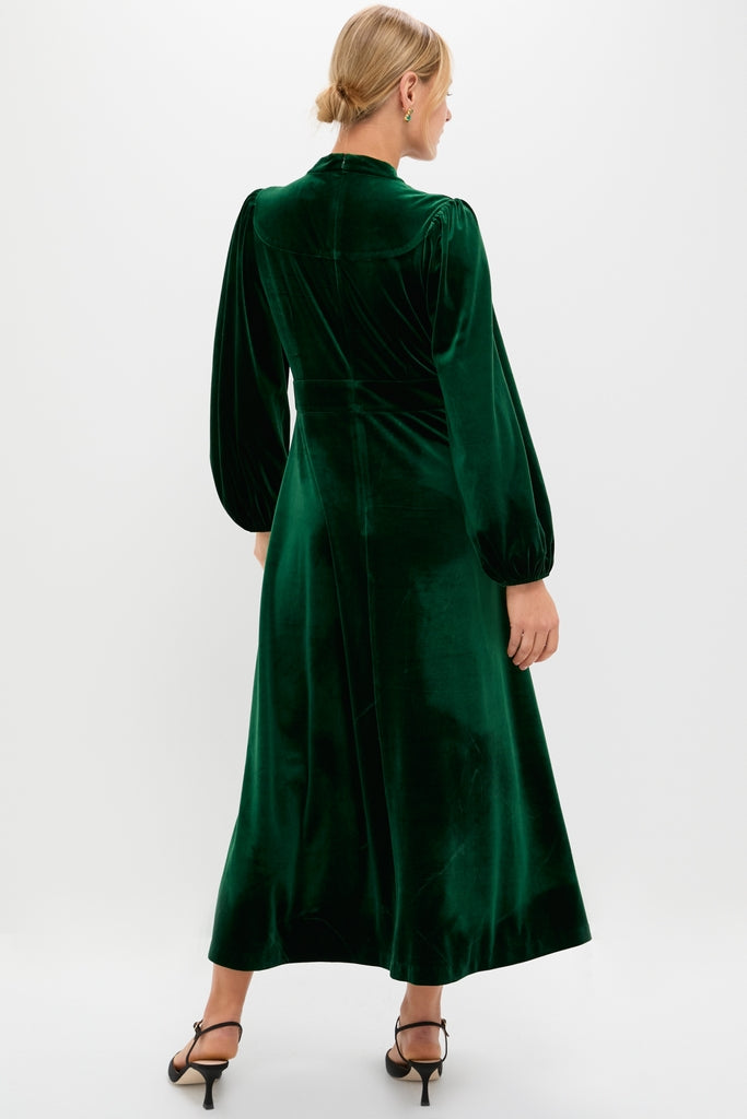 Woman in classic Green Velvet Serena Dress, featuring long balloon sleeves and fitted waist. Effortless elegance with high-end details, styled with black-heeled sandals. Perfect for fun, celebratory events.