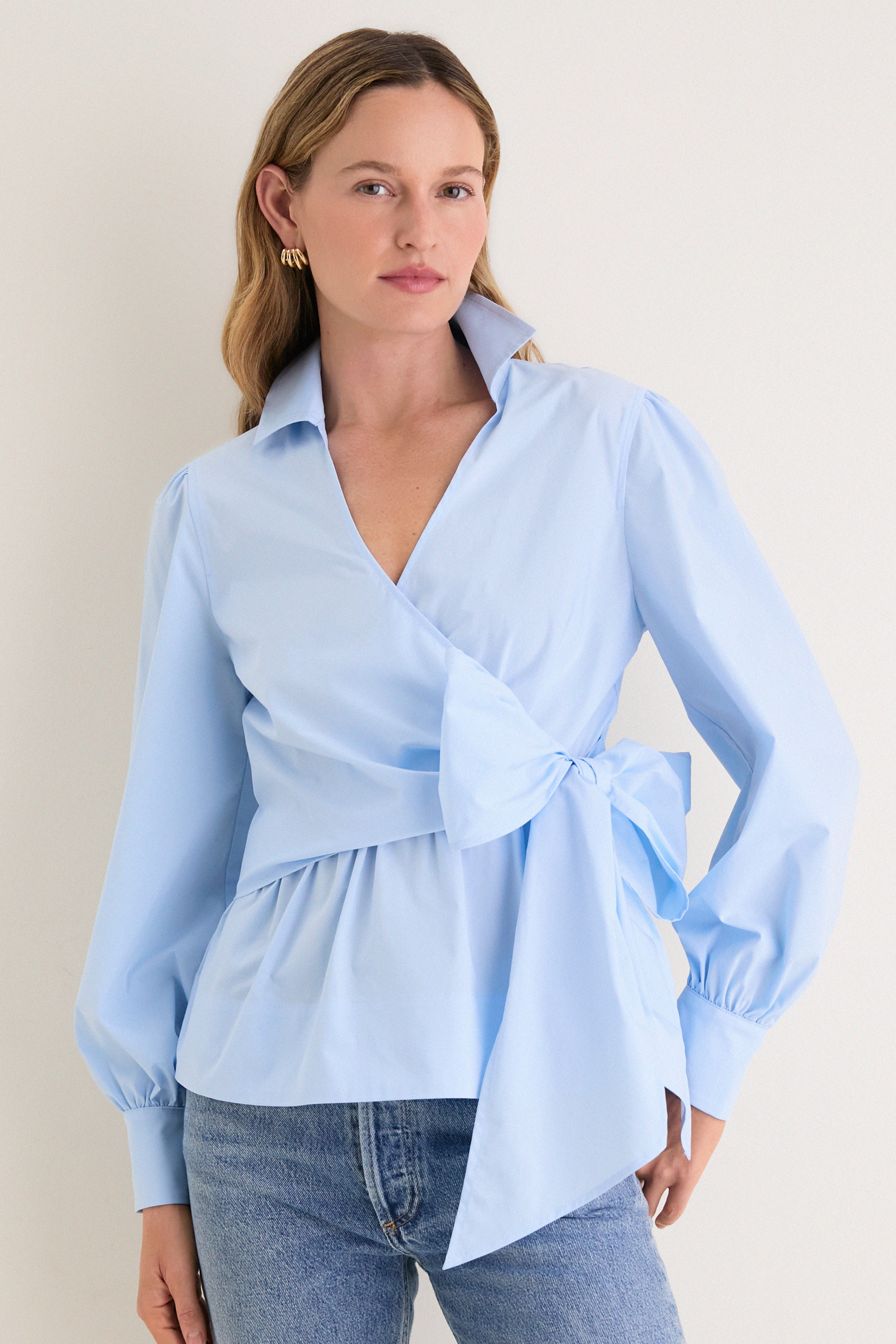 Female model in a classic, effortless French Blue Tie Front Bow Bea Blouse, showcasing its chic wrap design, peplum waist, and elegant self-tie bow.
