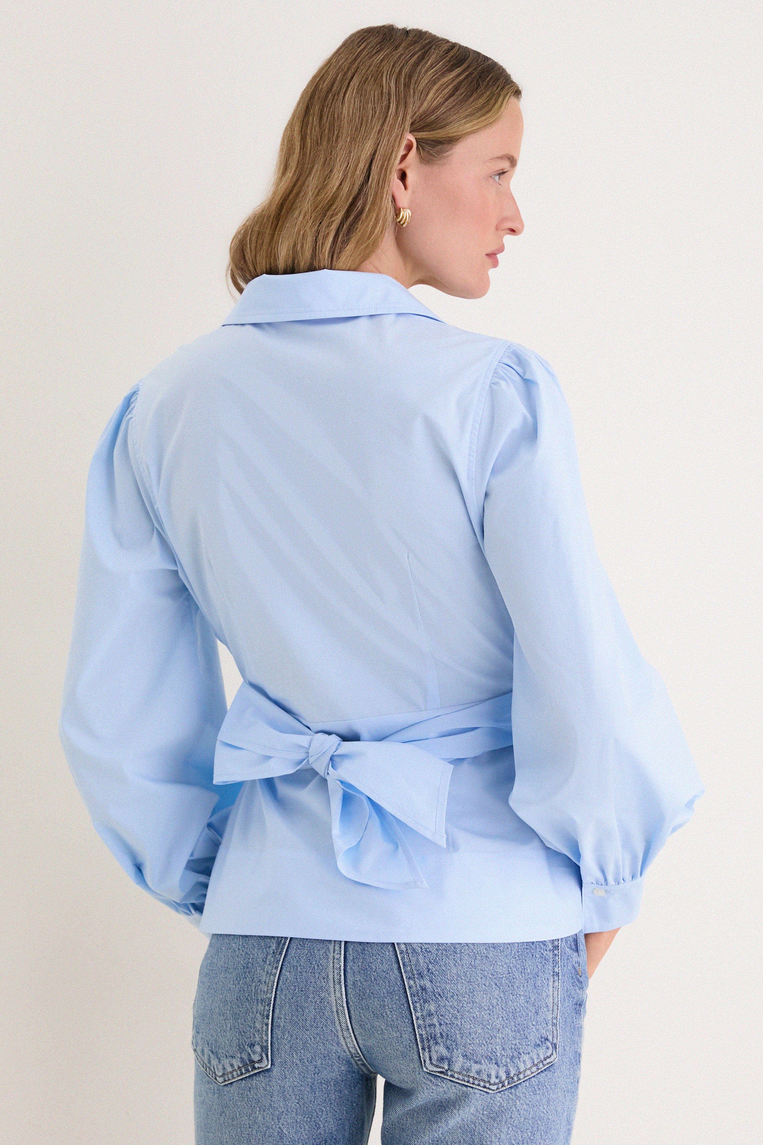 Back view of a person wearing the French Blue Tie Front Bow Bea Blouse, featuring a classic wrap design with a chic bow and slightly puffed sleeves.