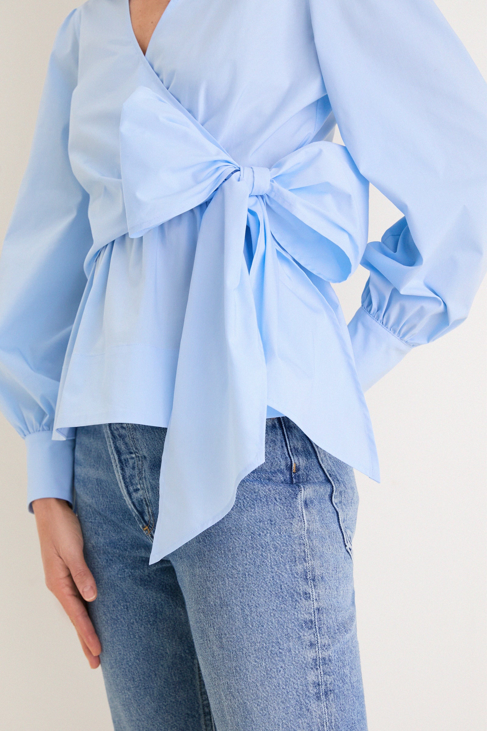 French Blue Tie Front Bow Bea Blouse with classic wrap design, effortless peplum waist, and fun oversized bow, paired stylishly with medium-wash jeans for a high-end look.