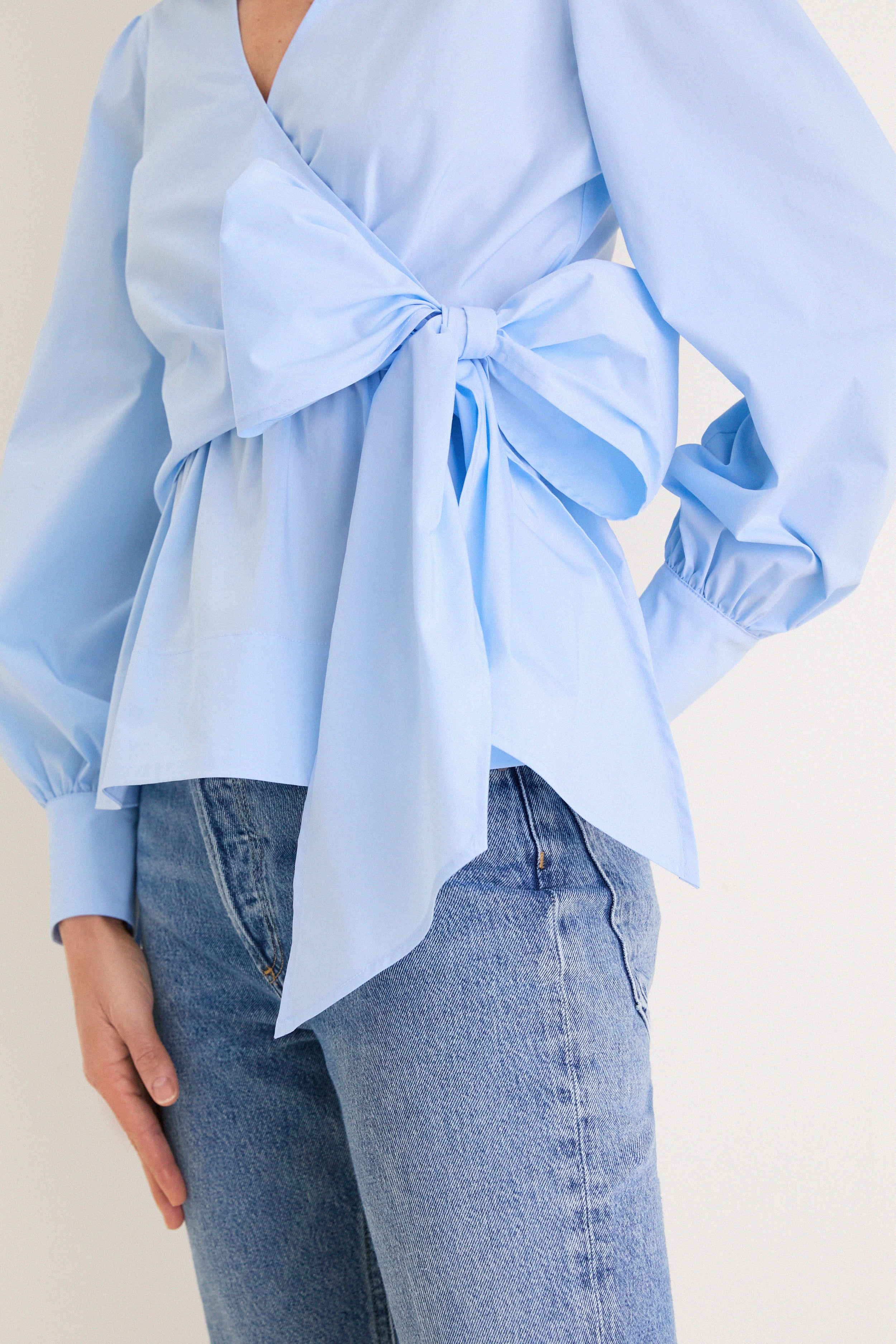 French Blue Tie Front Bow Bea Blouse with classic wrap design, effortless peplum waist, and fun oversized bow, paired stylishly with medium-wash jeans for a high-end look.