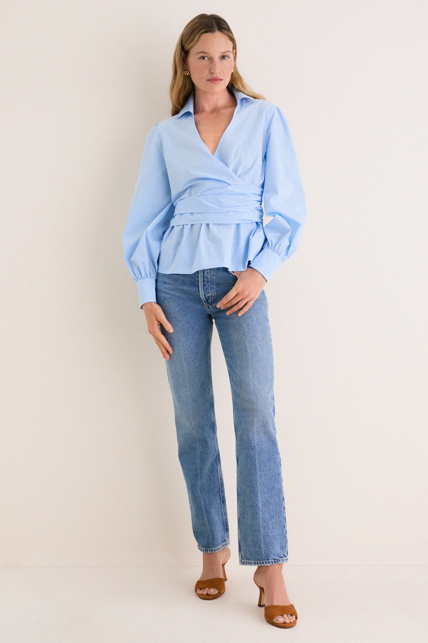 Woman models the classic French Blue Tie Front Bow Bea Blouse, featuring an effortless wrap style and peplum waist, paired with light jeans and tan mule heels.