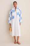 Woman wearing the Blue and White Quilted Cropped Townley Jacket, a classic, high-end layer with patchwork checks, styled effortlessly over a white dress, holding a woven handbag.
