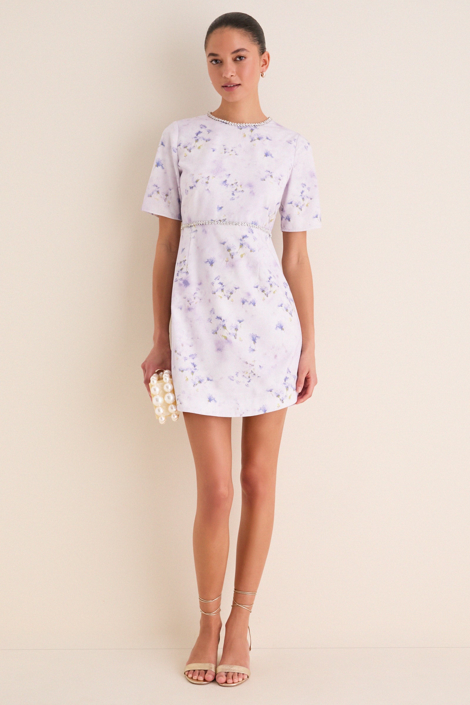 Woman in Lilac Floral Crystal Embellished Floria Mini Dress, clutch and sandals, exuding classic, effortless elegance and fun for elevated daytime events.
