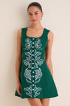 Young woman in a sleeveless, forest-green A-line dress with silver-tone buttons and white embroidery, embodying classic, effortless style and high-end fun.
