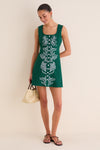Young woman in a forest-green mini dress with white embroidery and buttons, holding a straw tote, embodying classic, effortless, and fun style from Tuckernuck.