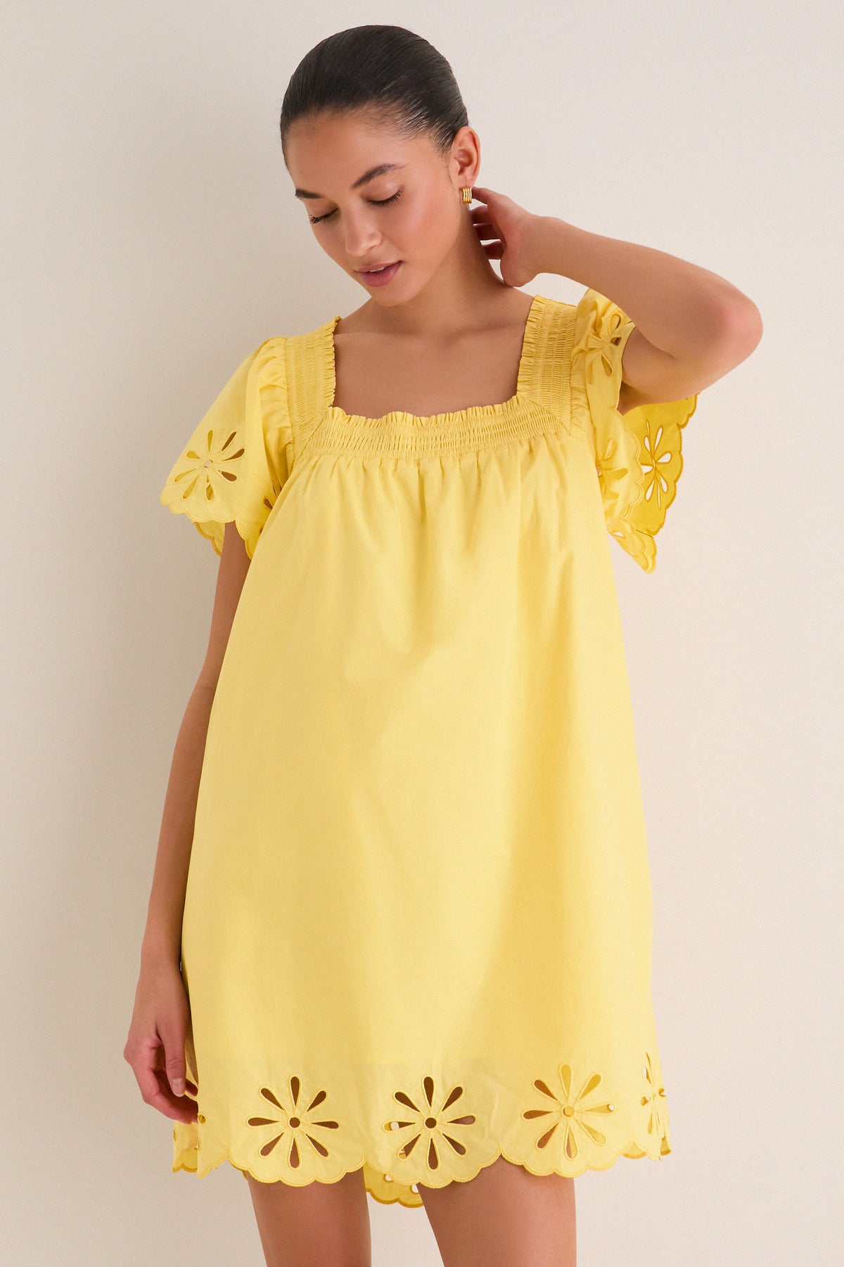 Model in a classic, bright yellow babydoll mini dress with a square neckline, flutter sleeves, and eyelet detailing; an effortless and fun high-end fashion piece.