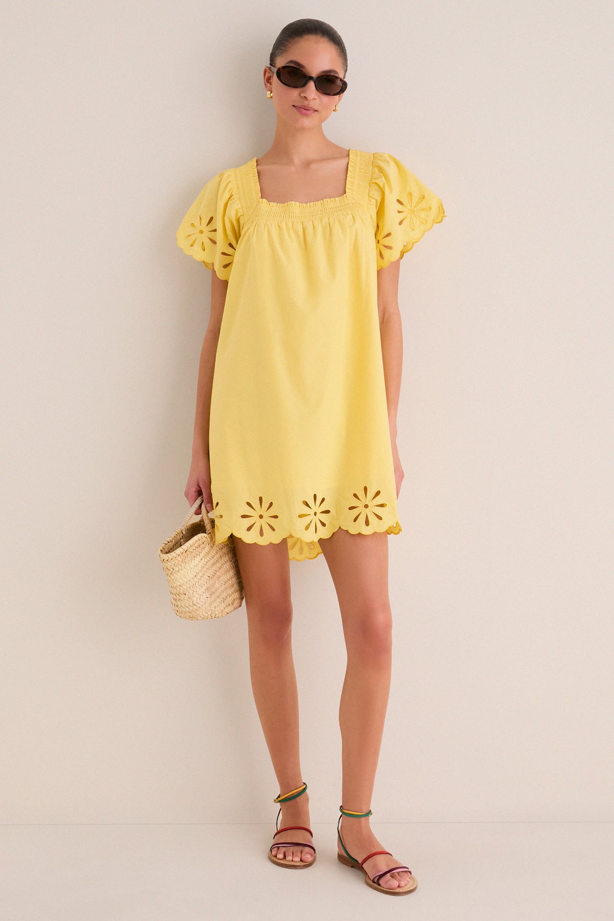 Woman in a classic, yellow Floral Eyelet Daisy Mini Dress with puffed sleeves, scalloped hem, paired with cat-eye sunglasses and a straw bag, exuding effortless, fun style.