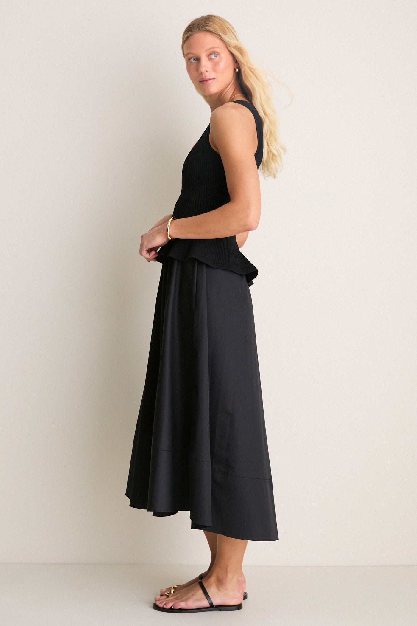 Model in Black Peplum Cara Dress, a classic, effortless ensemble with a form-fitting top and flowing midi skirt, styled with gold accessories and flat sandals for a high-end, fun look.