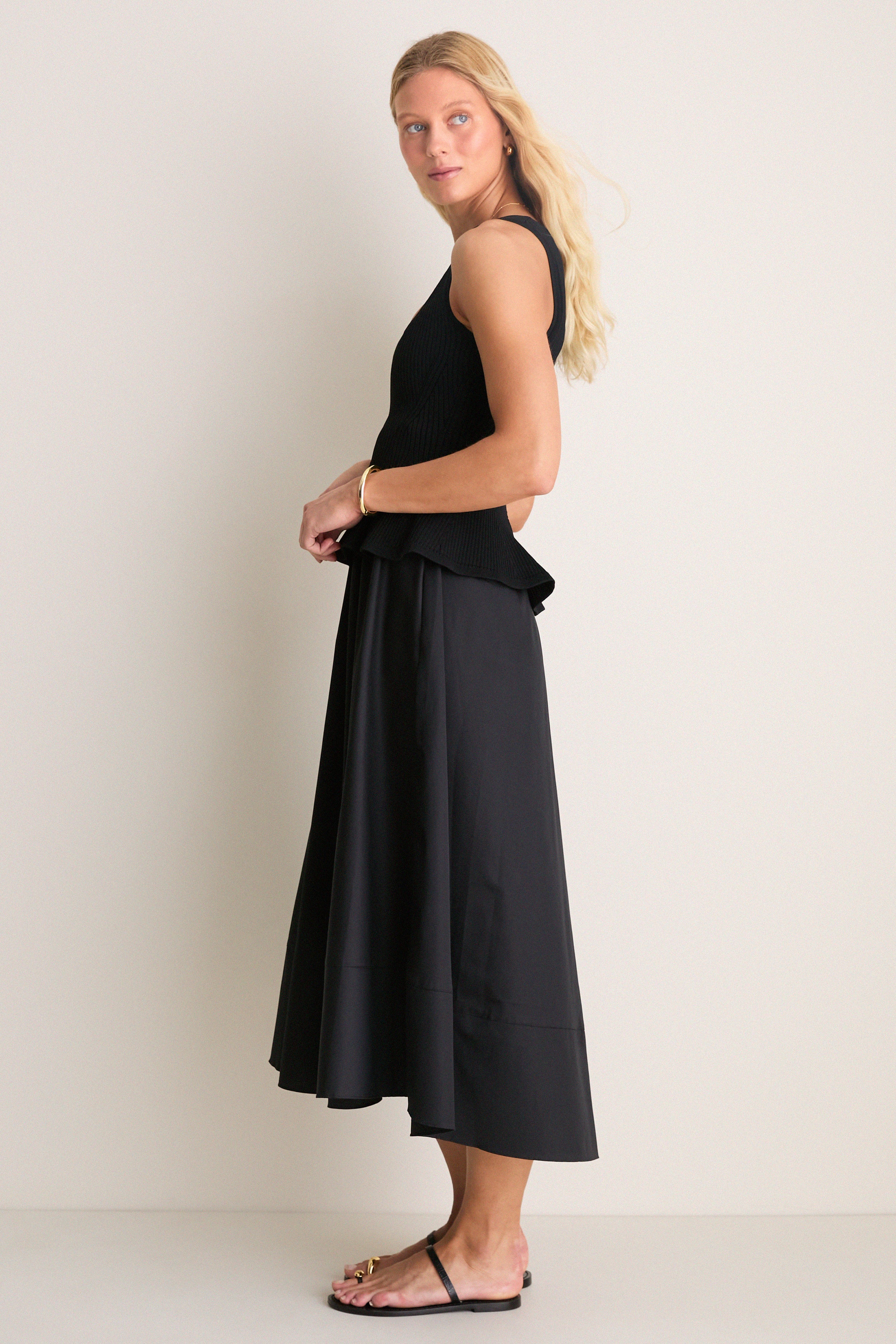 Model in Black Peplum Cara Dress, a classic, effortless ensemble with a form-fitting top and flowing midi skirt, styled with gold accessories and flat sandals for a high-end, fun look.