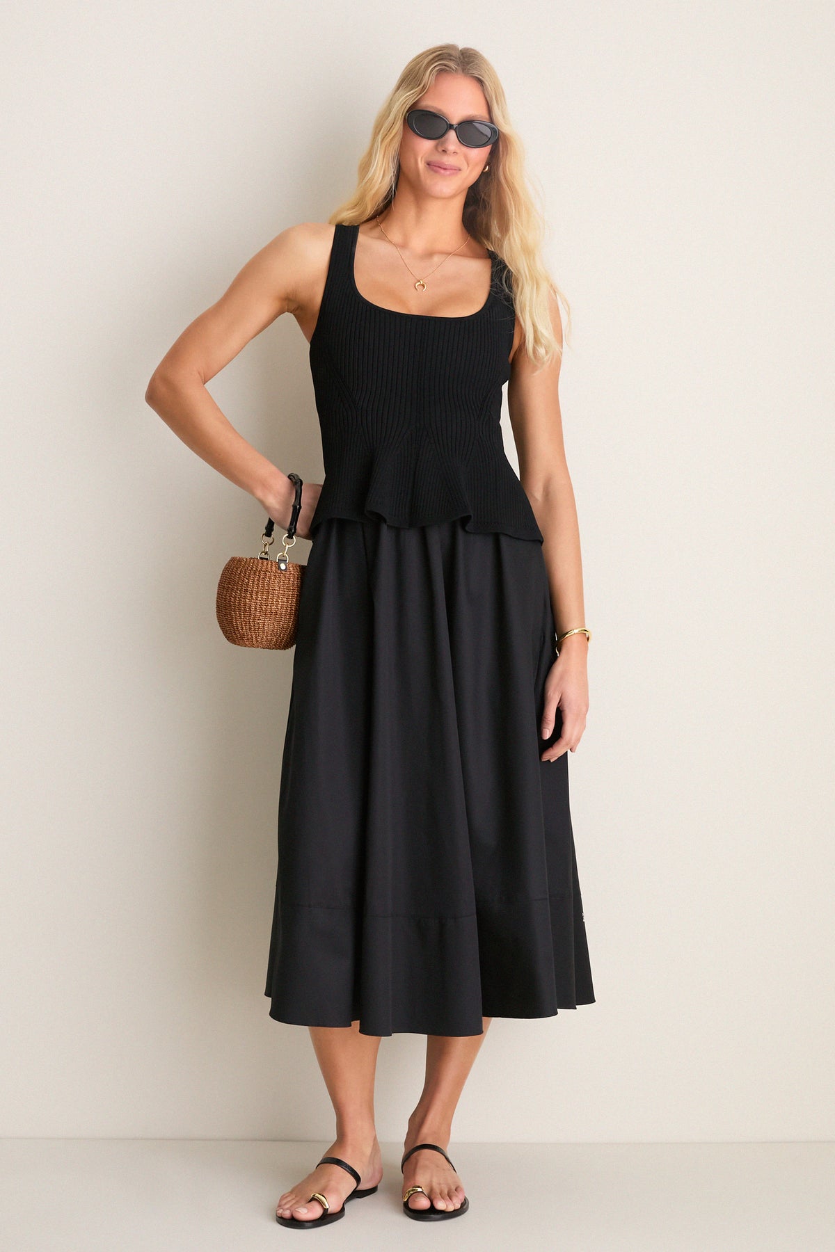 Young woman in a Black Peplum Cara Dress, featuring a rib-knit top and flared skirt, accessorized with black sandals, sunglasses, and a straw handbag. Classic and effortlessly chic.
