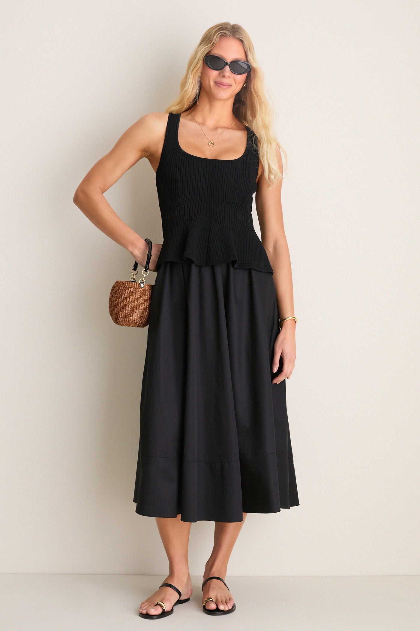 Young woman in a Black Peplum Cara Dress, featuring a rib-knit top and flared skirt, accessorized with black sandals, sunglasses, and a straw handbag. Classic and effortlessly chic.