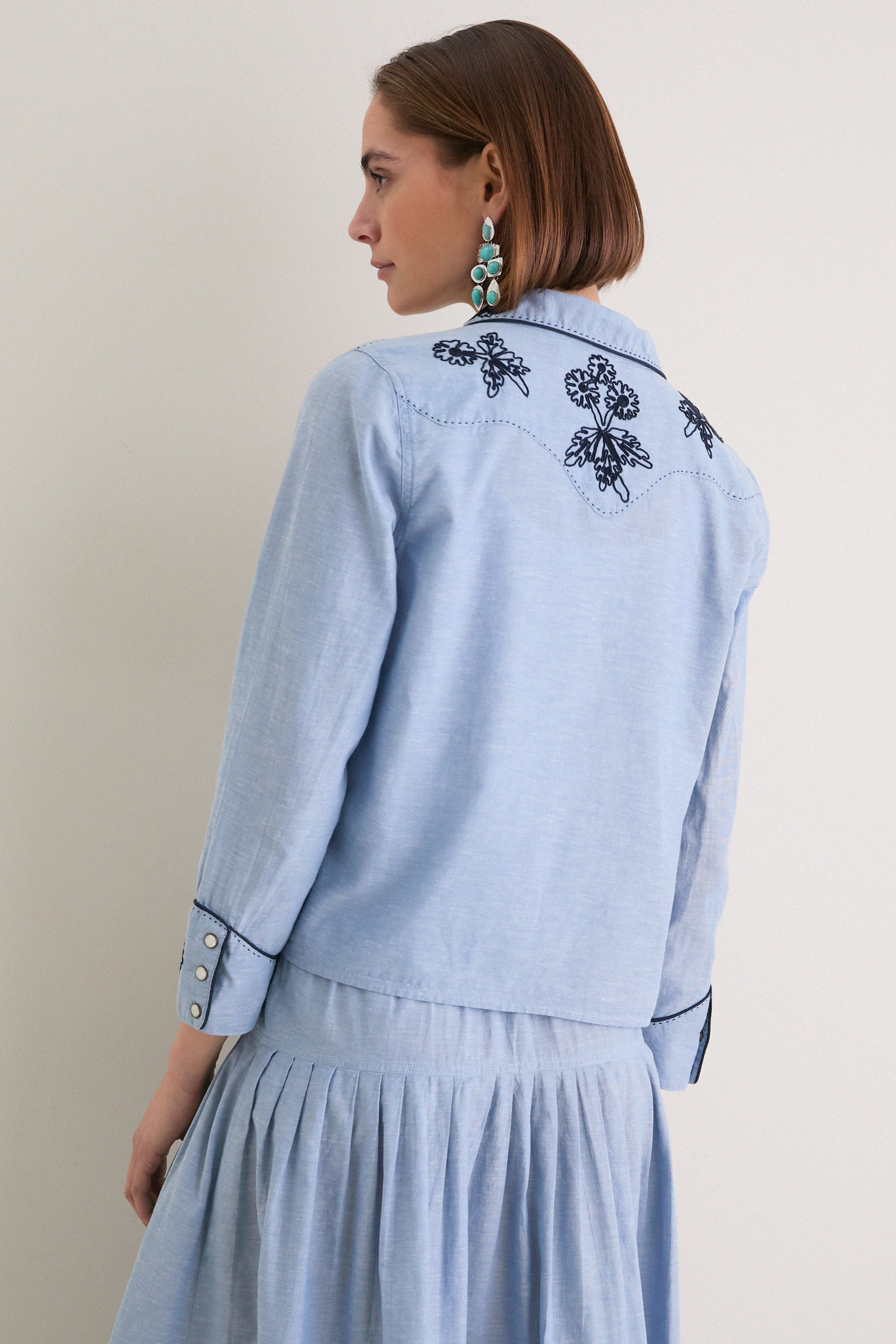Woman in profile wearing the Chambray Embroidered Western Shirt; classic, effortless style with dark embroidery, snap-button cuffs, and a relaxed fit.