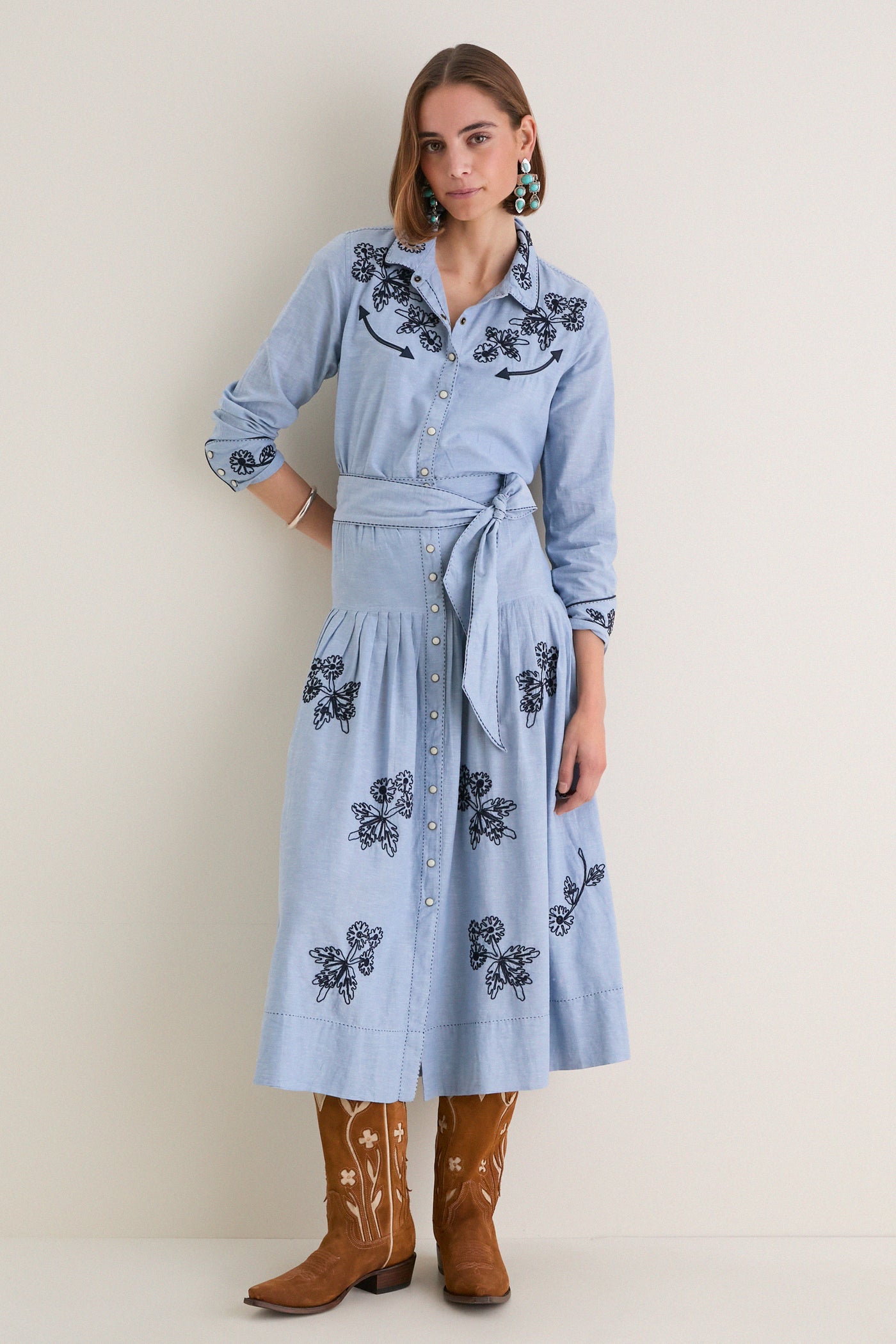 Woman models The Chambray Embroidered Western Shirt with floral embroidery, rolled sleeves, and a self-fabric belt, paired with turquoise earrings and cowboy boots, embodying classic, effortless style.