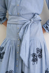 The Chambray Embroidered Highland Skirt features a classic, effortless design with floral embroidery, a self-tie sash, pleats, and fun snap buttons for a high-end look.