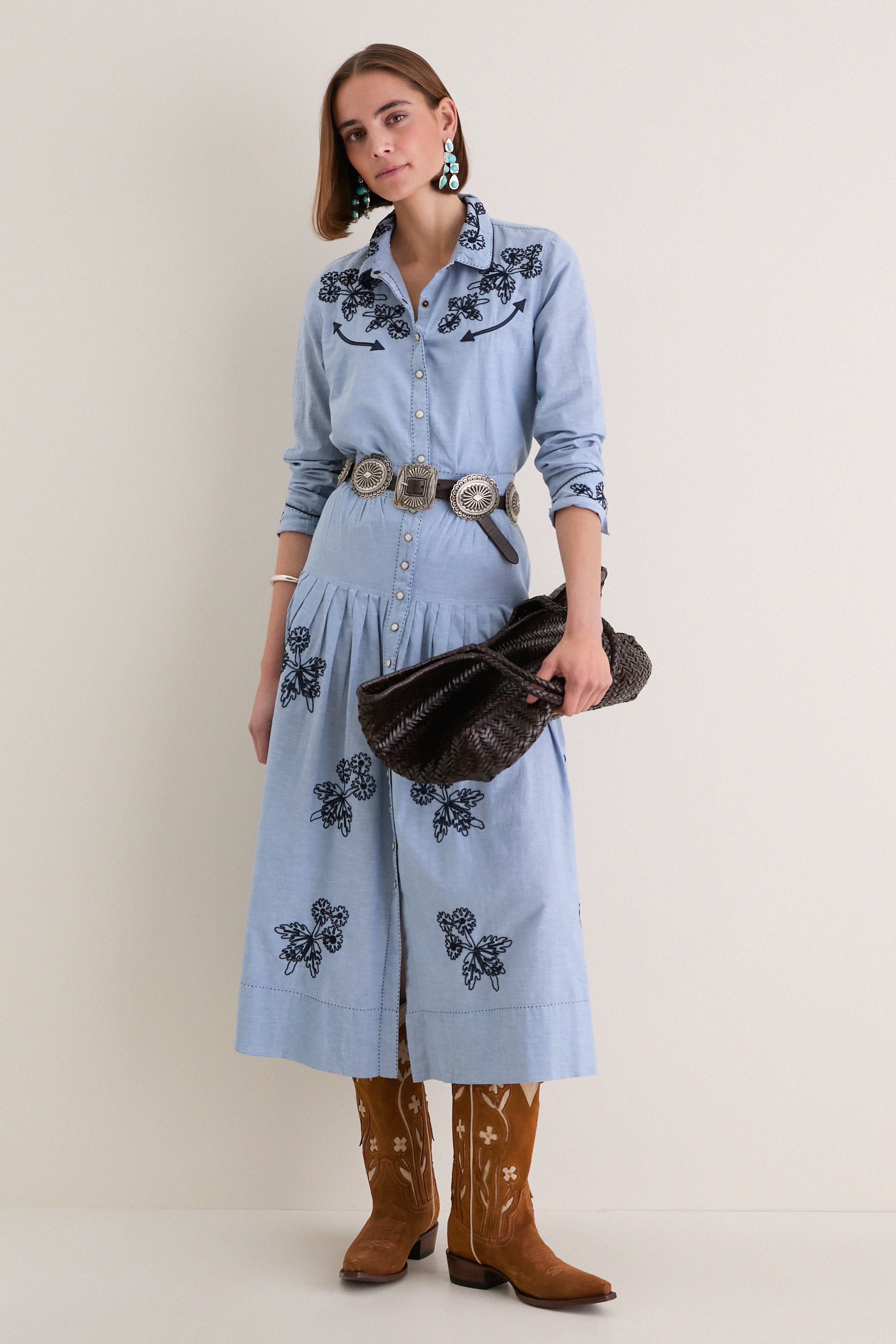 Woman wearing The Chambray Embroidered Highland Skirt with navy floral embroidery, paired with a silver concho belt, turquoise earrings, and cowboy boots for a classic, effortless look.