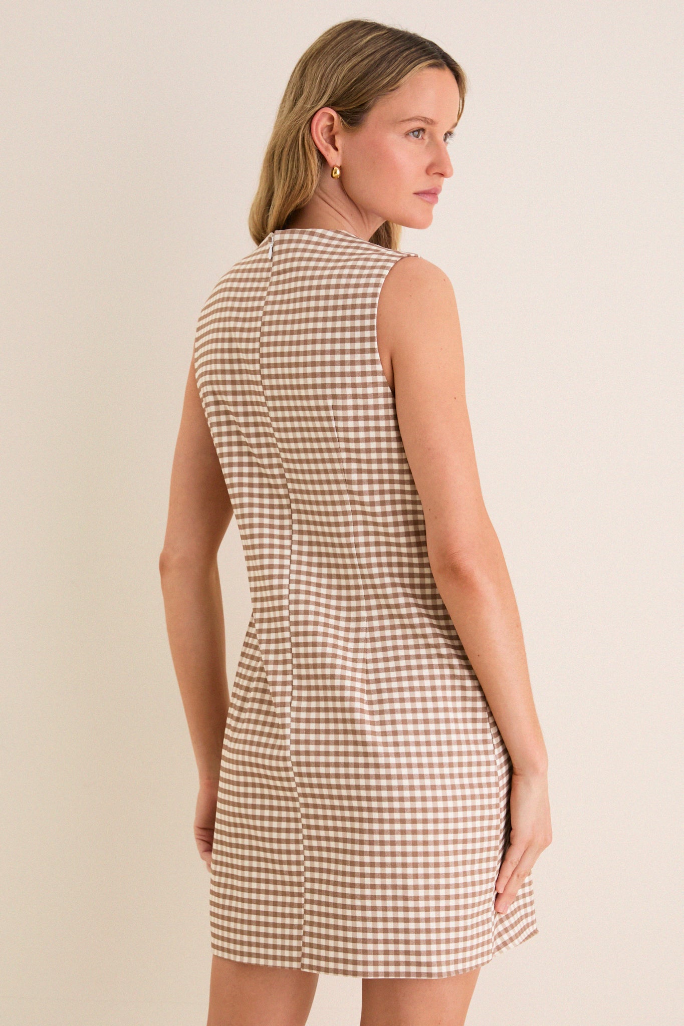 Woman modeling the Mocha Gingham Yvette Mini Dress, showcasing its classic, sleeveless design and effortless vibe, perfectly embodying Tuckernuck's fun and high-end style.