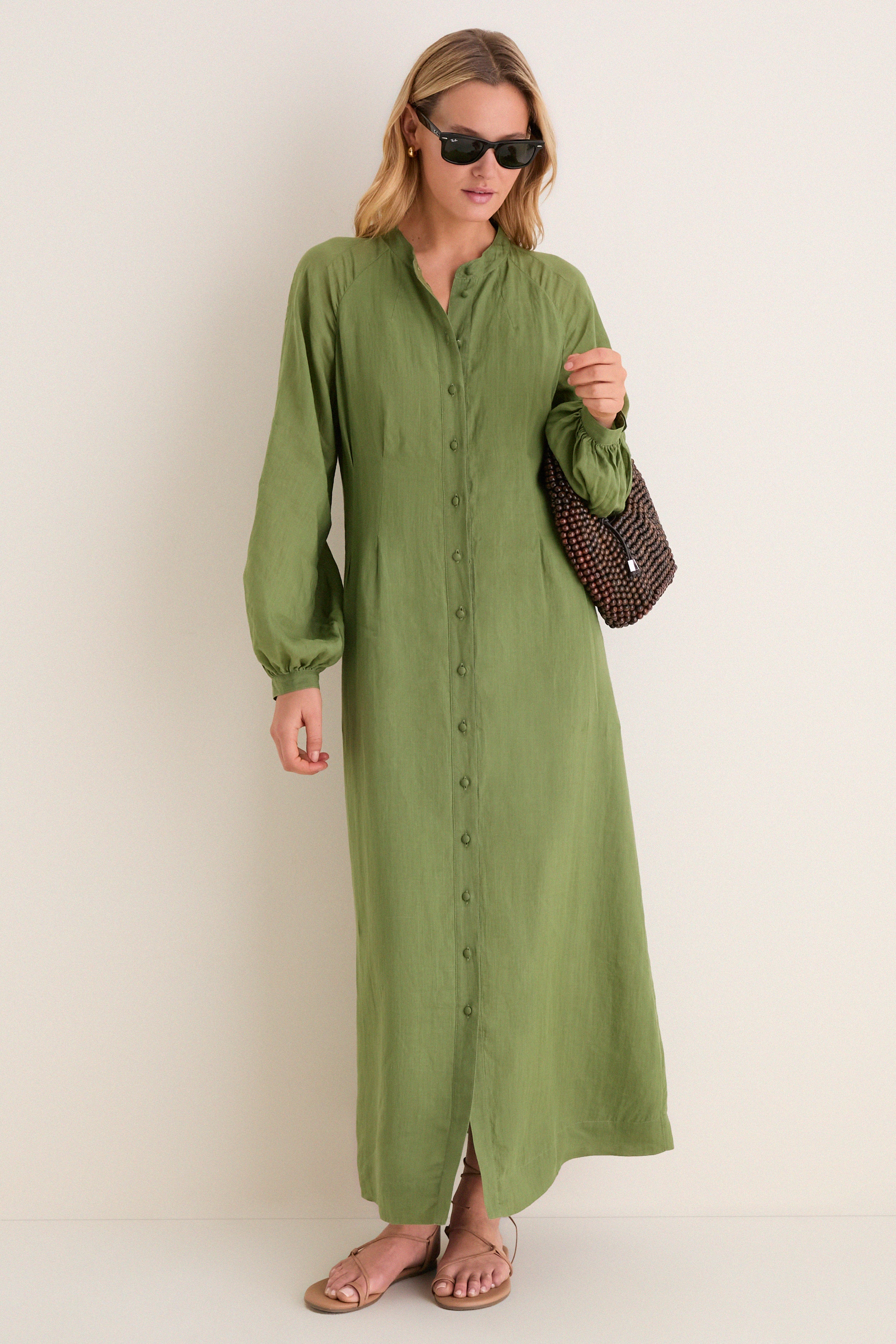 Woman wearing Cedar Green Button Down Granier Dress with sunglasses, holding a dark brown handbag. The classic, high-end dress features a relaxed fit and removable belt, paired with tan sandals.