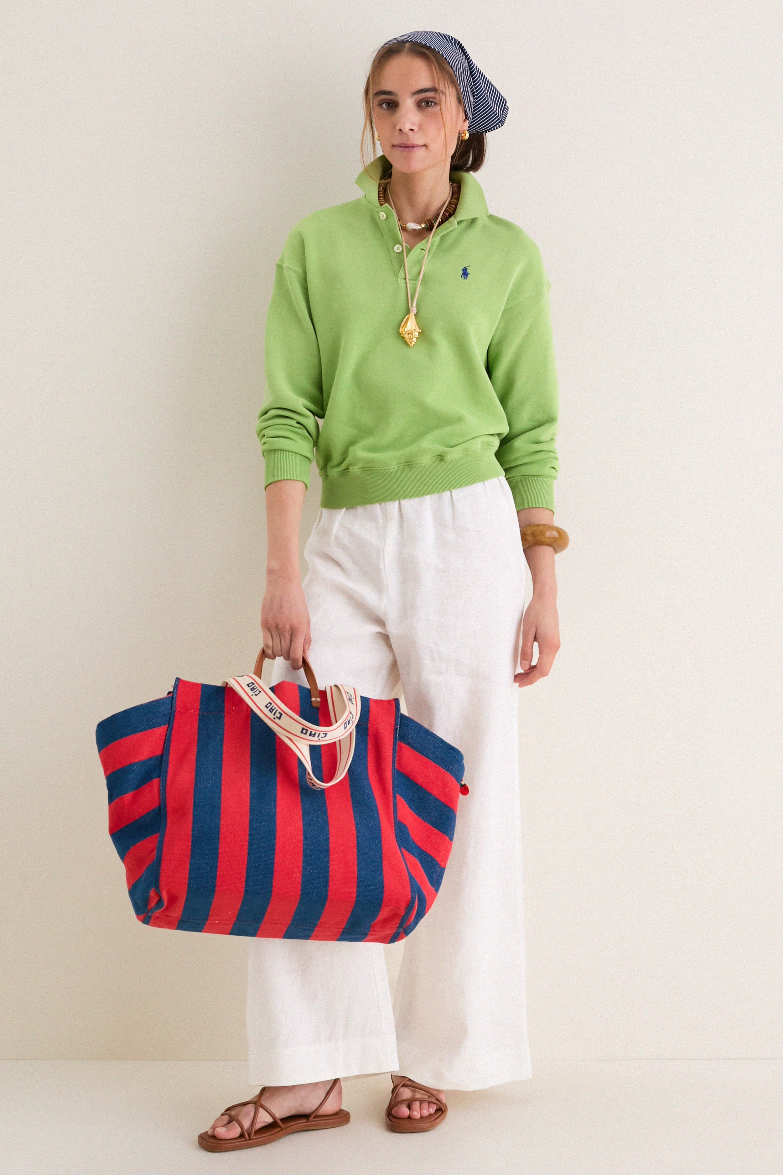 A person styled in a relaxed, summery look holds the Poppy and French Blue Beach Tote, showcasing its classic, effortless design and fun, high-end stripes.