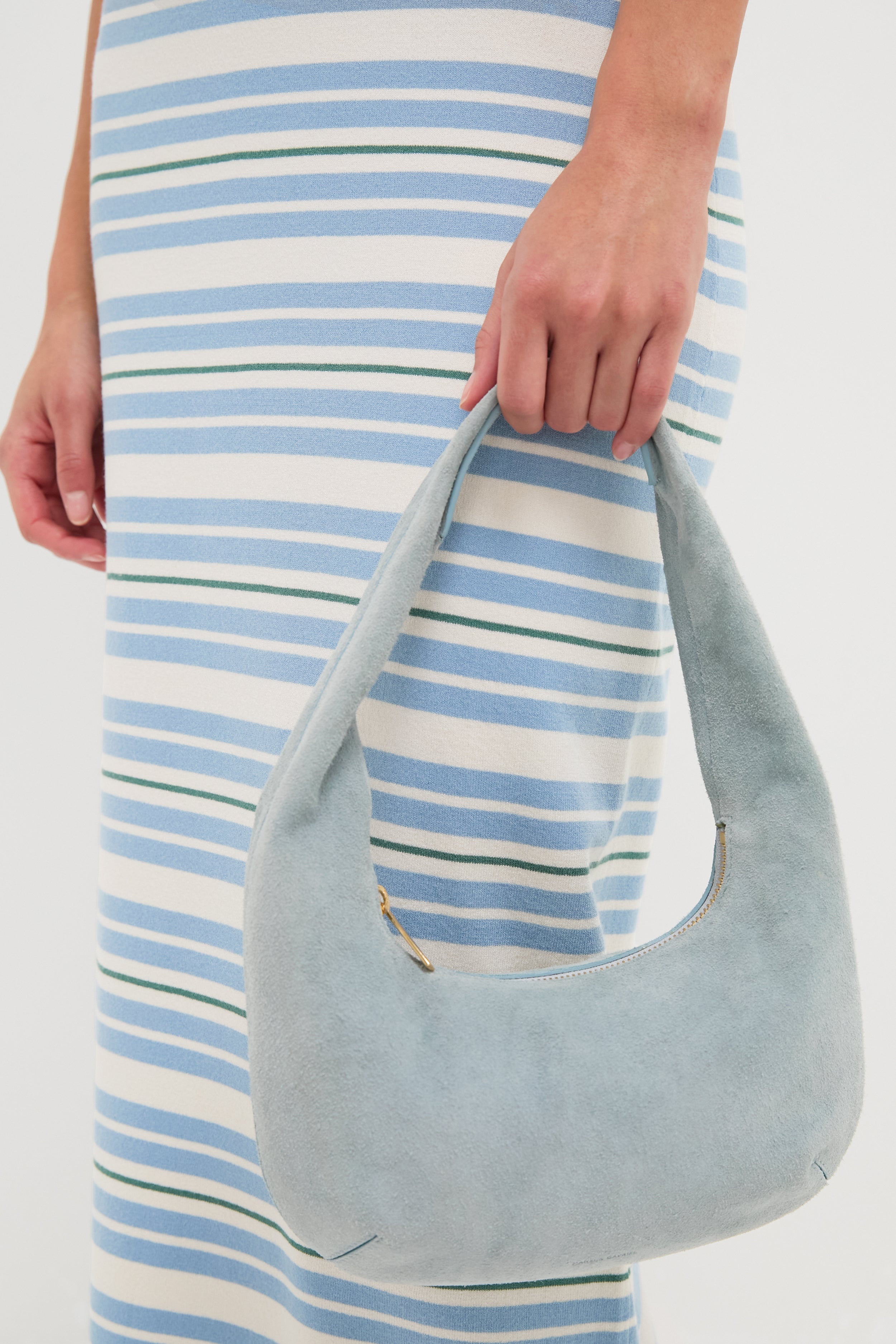 Person holding the Baby Blue Gnocchi Bag, a classic, crescent-shaped suede hobo with zip closure, effortlessly complementing a form-fitting, striped dress for a fun, high-end look.