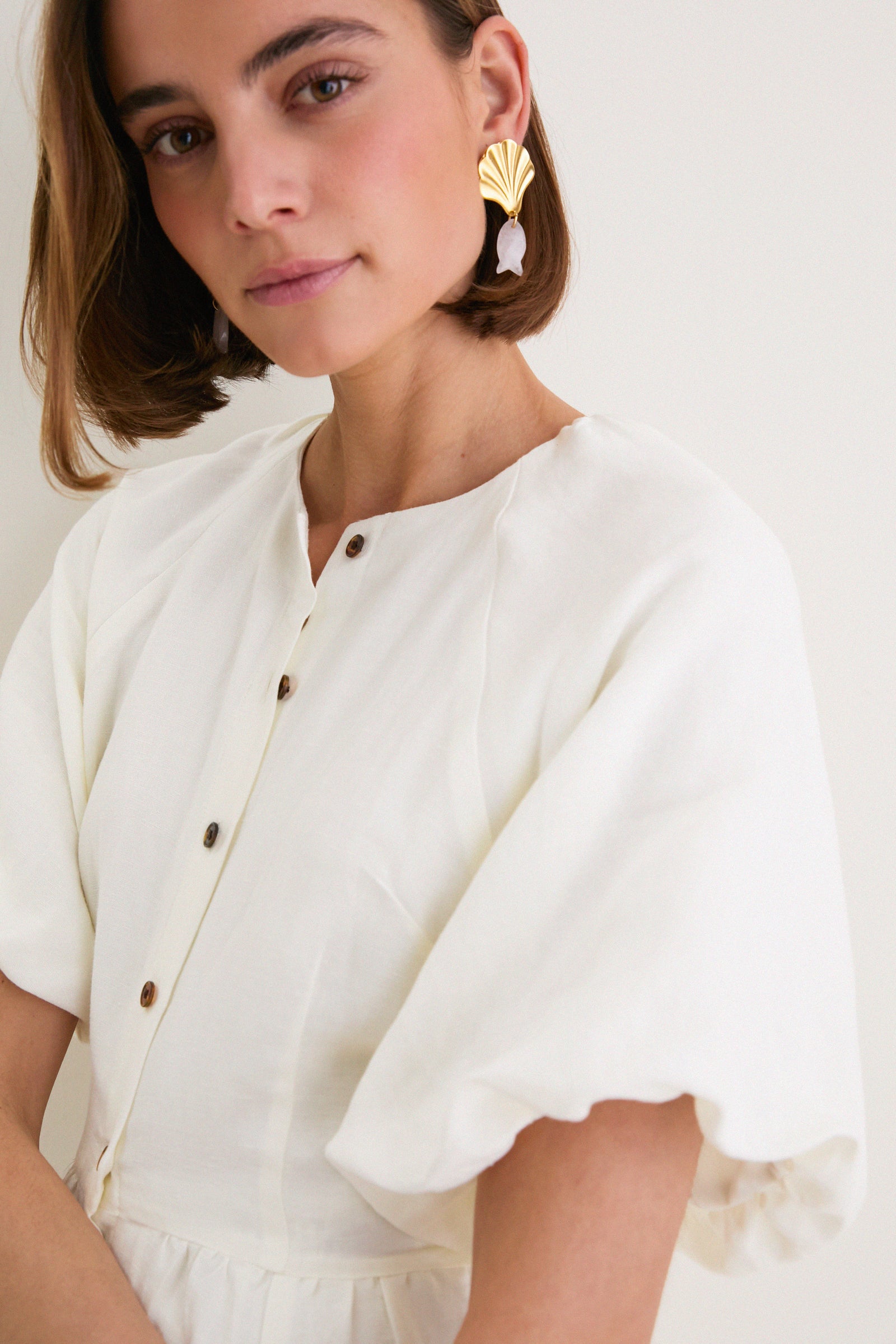 Young woman in classic Star White Bastille Short Dress with voluminous sleeves and high neckline, exuding effortless elegance. She wears a chic gold scallop-shell drop earring.